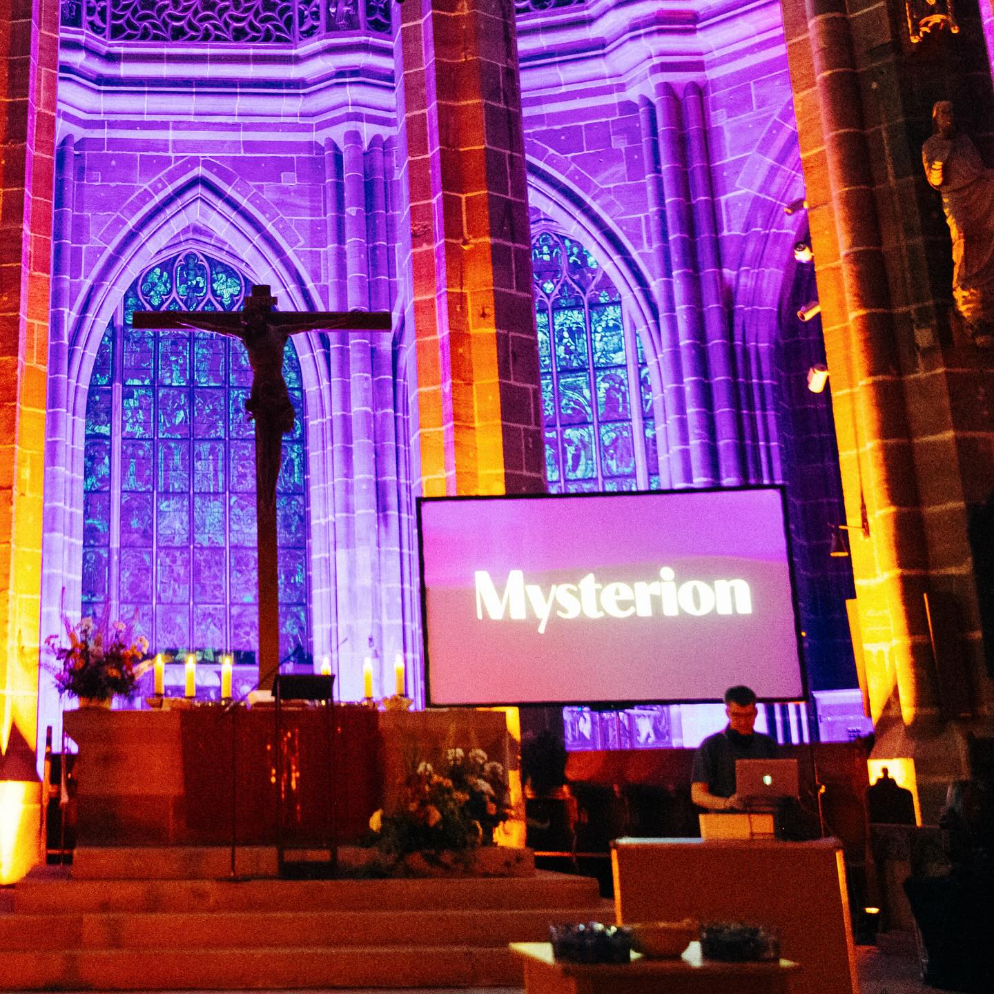 Eine alte Kirche in pinkes Licht getaucht. Auf einem großen Bildschirm steht "Mysterion". Davor legt ein DJ auf.